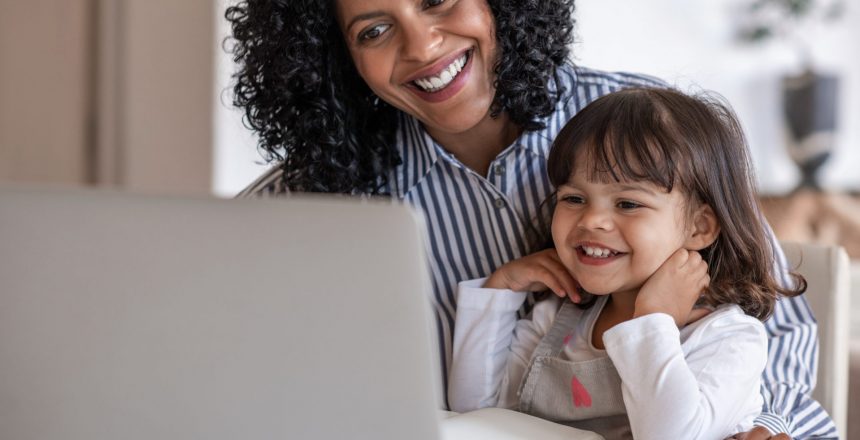 Busy mother and entrepreneur smiling while working on a laptop with her cute little girl sitting on her lap